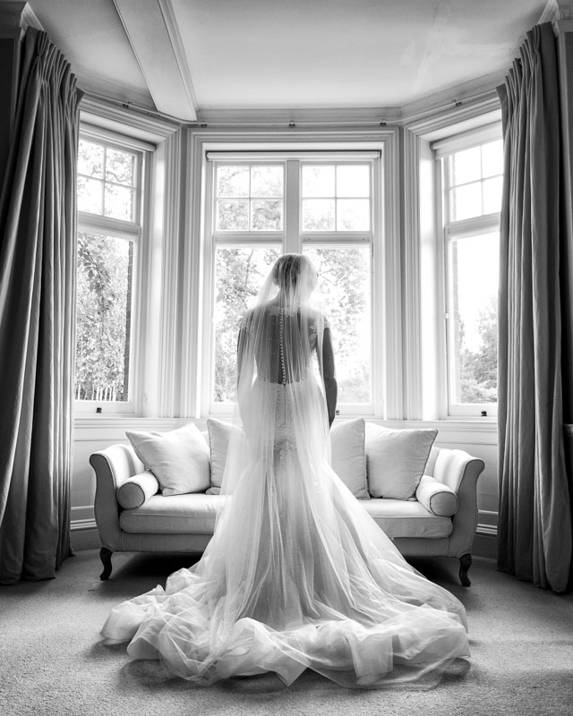 Black and white portrait of bride in beautiful flowing dress in front of window at Chippenham Park in Cambridgeshire